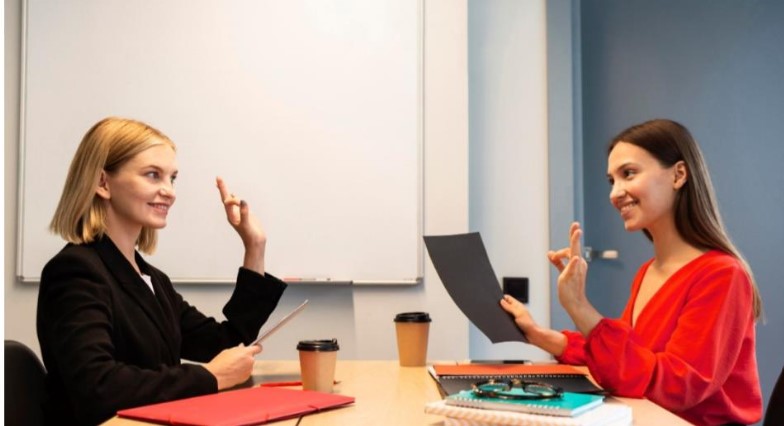 Grafika dekoracyjna- dwie kobiety siedzące w biurze rozmawiające w języku migowym