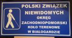 tablica informacyjna na niebieskim tle napis Polski Związek Niewidomych Okręg Zachodniopomorski Koło Terenowe w Białogardzie
