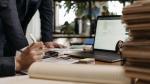 Biurko z dokumentami i laptopem, dłonie dwóch osób, jedna z osób trzyma długopis i kalkulator