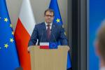 Ubrany w garnitur minister Marcin Łoboda stoi za mównicą. W tle flagi Polski i Unii Europejskiej.