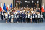 Duża grupa osób stoi na scenie i prezentuje oprawione certyfikaty podczas oficjalnej uroczystości, na tle flag Polski i Unii Europejskiej oraz ściany z logo instytucji