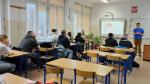 Trener stoi w rogu sali i prowadzi zajęcia. Obok wyświetlona prezentacja. Uczniowie siedzą w ławkach szkolnych i patrzą na trenera