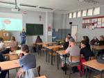 Trenerka stoi w klasie i prowadzi zajęcia. Obok wyświetlona prezentacja. Uczniowie siedzą w ławkach szkolnych i patrzą na trenerkę